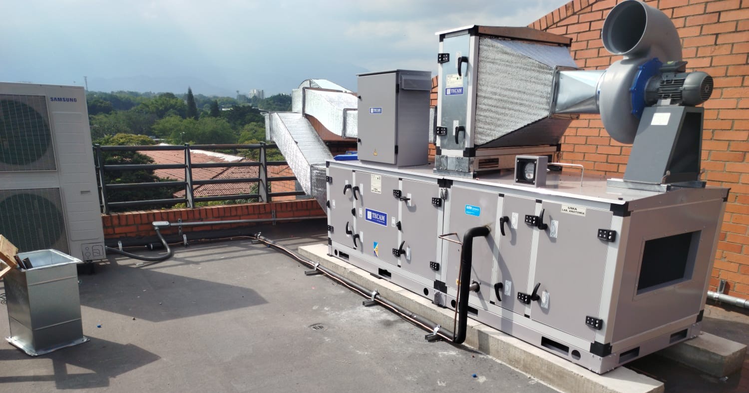 Large industrial air conditioning unit installed on a rooftop, surrounded by ventilation ducts and a brick wall. The unit is stationary and appears newly installed. The wider environment includes a clear sky, distant trees, and nearby buildings, creating a professional and functional atmosphere. No visible text in the image.